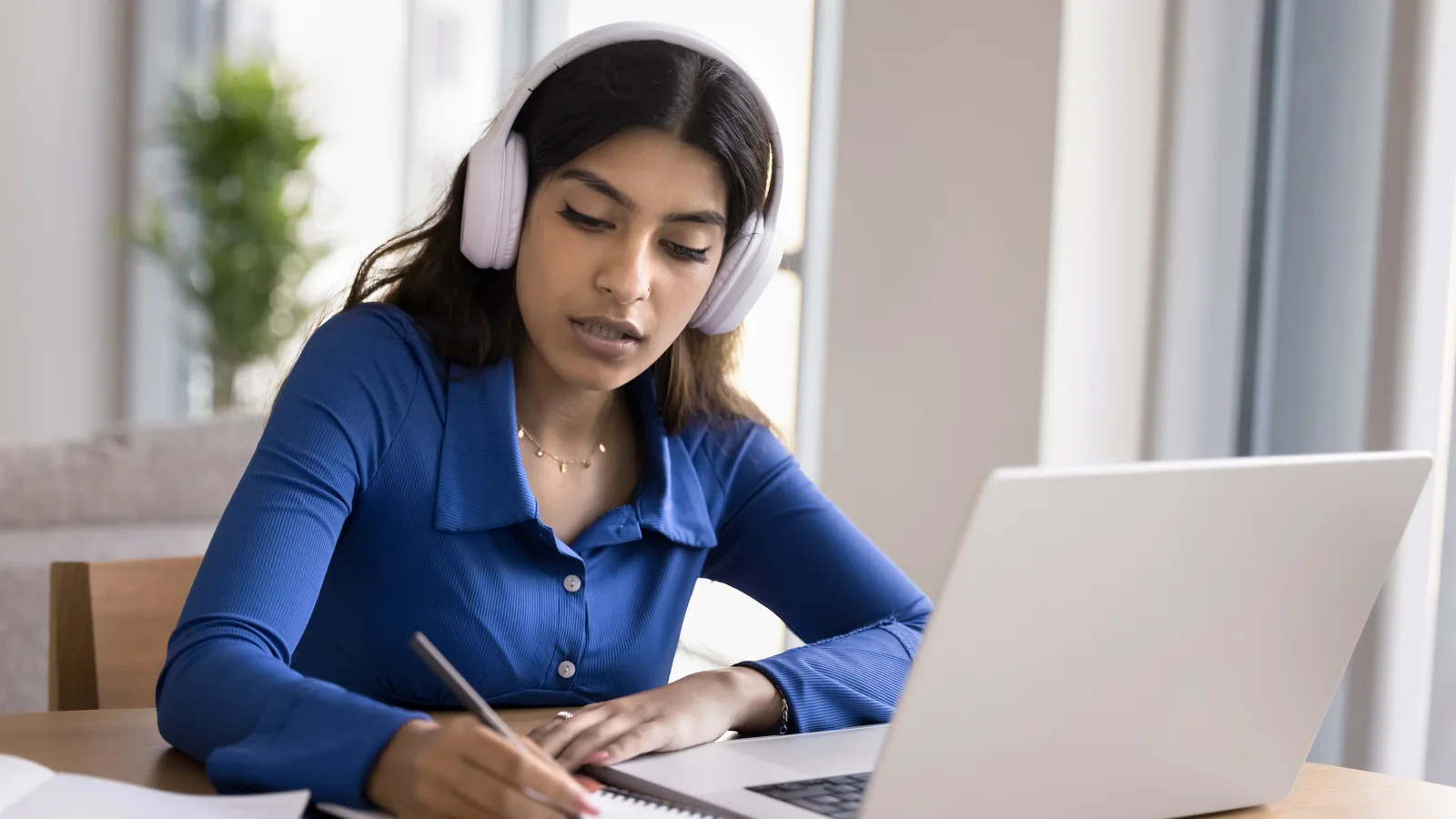 Une femme portant un casque prend des notes dans un cahier devant un ordinateur portable.