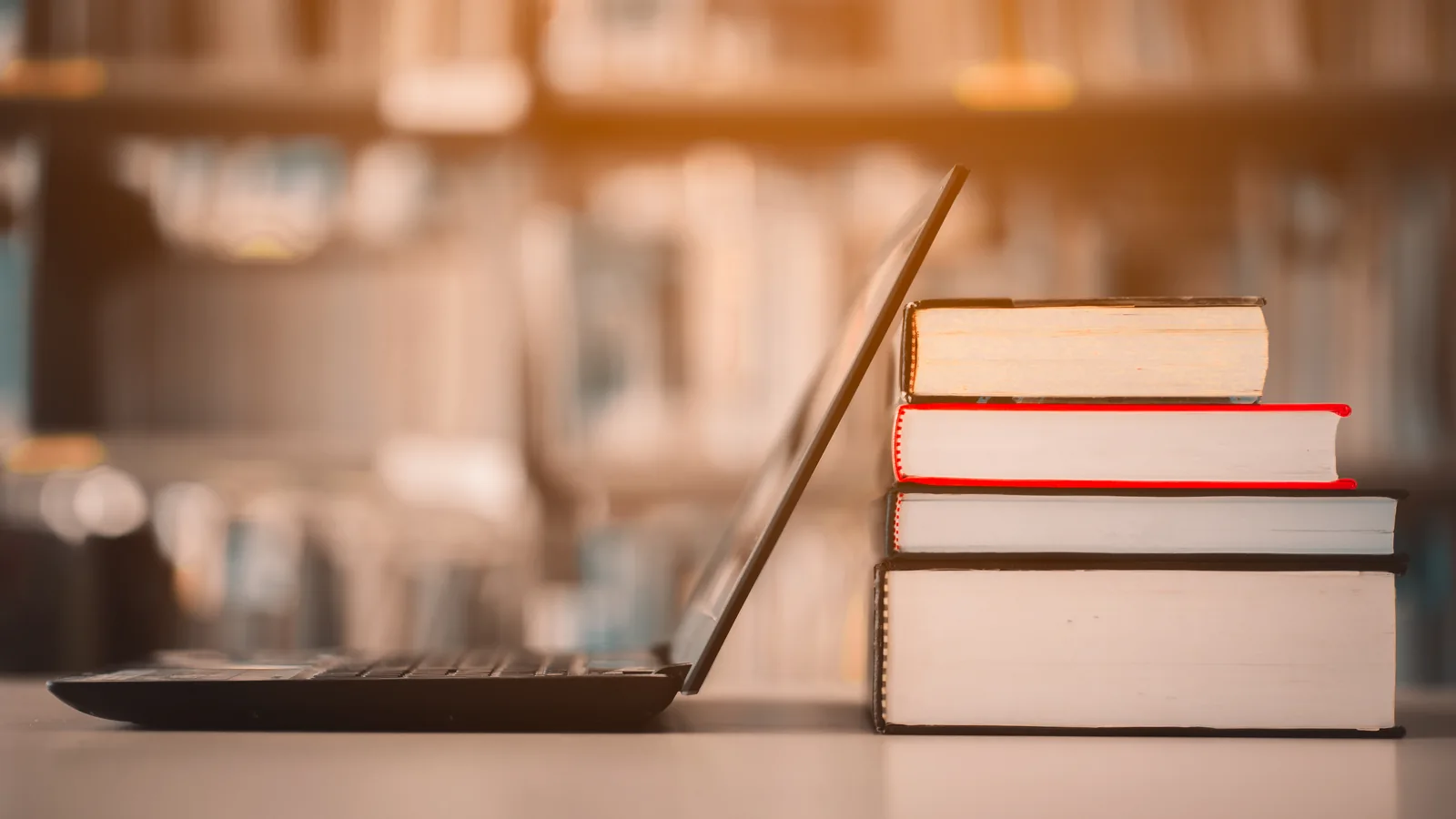 Un ordinateur portable ouvert à côté d’une pile de livres, devant les rayonnages d’une bibliothèque.
