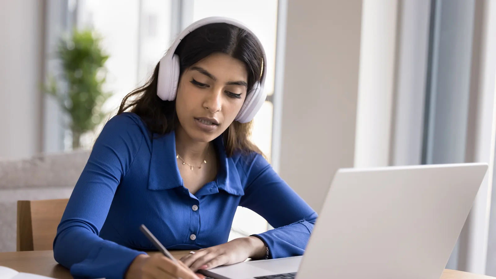 Une personne prend des notes dans un carnet devant un ordinateur portable, un casque sur les oreilles.