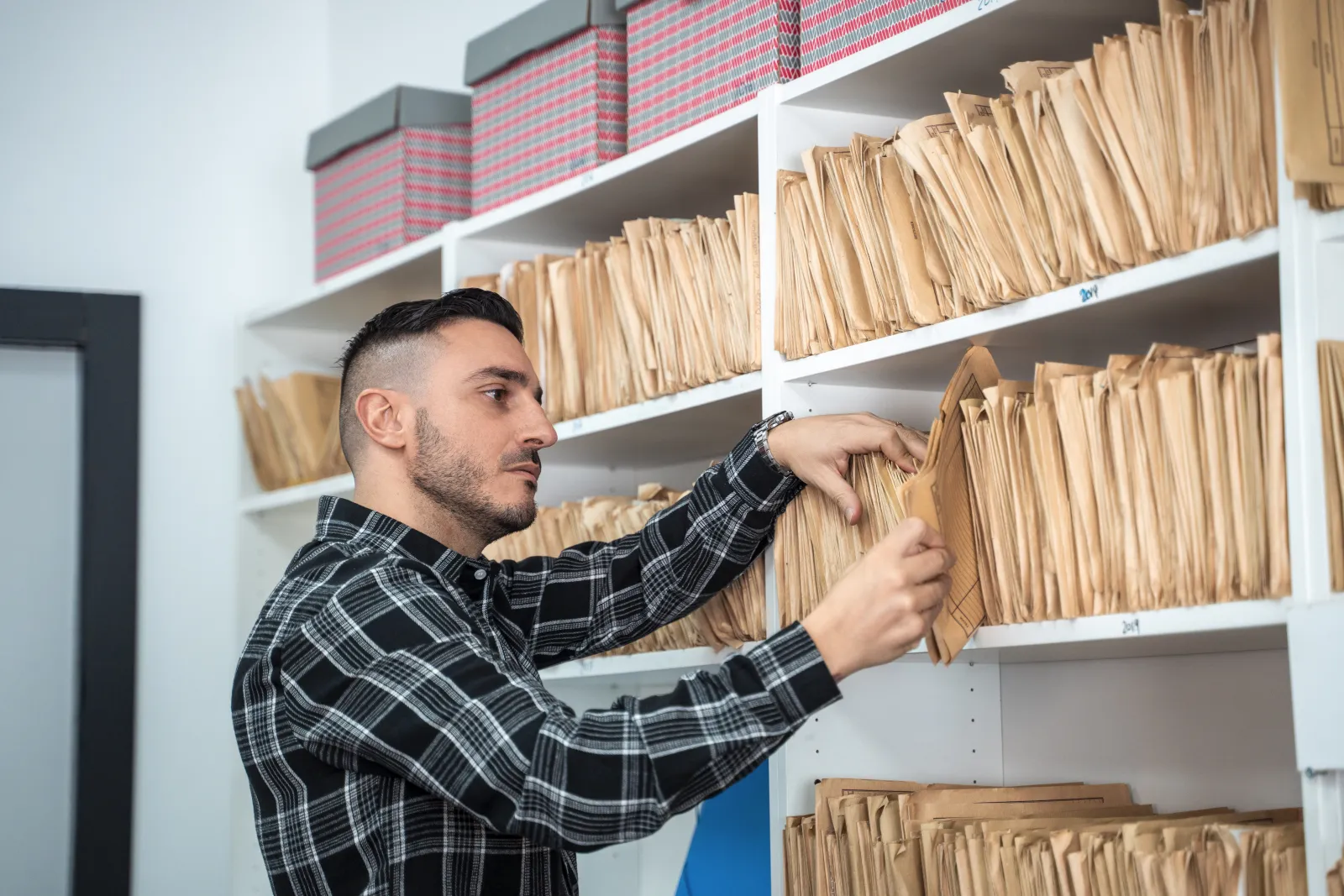 Un professionnel consulte des dossiers papier rangés sur les étagères d’une salle d’archives.