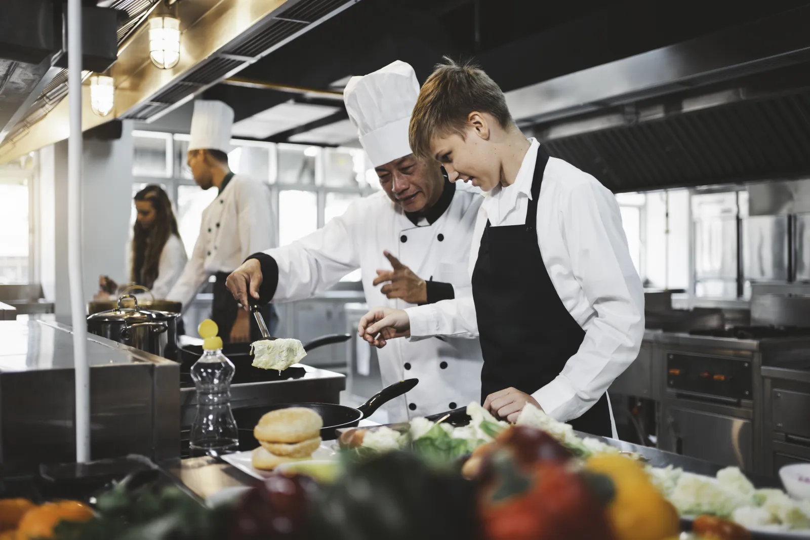 Un chef montre un geste de cuisine à un élève devant une plaque de cuisson.