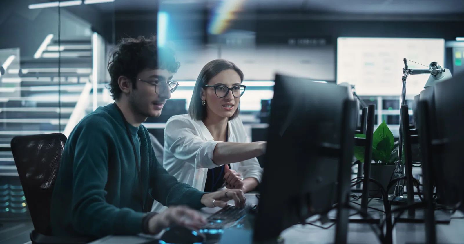 Deux collègues échangent devant les écrans de leur poste informatique.