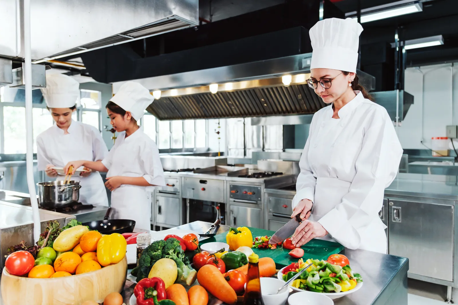 Des personnes en tenue de cuisine préparent des légumes et des plats dans une cuisine professionnelle.