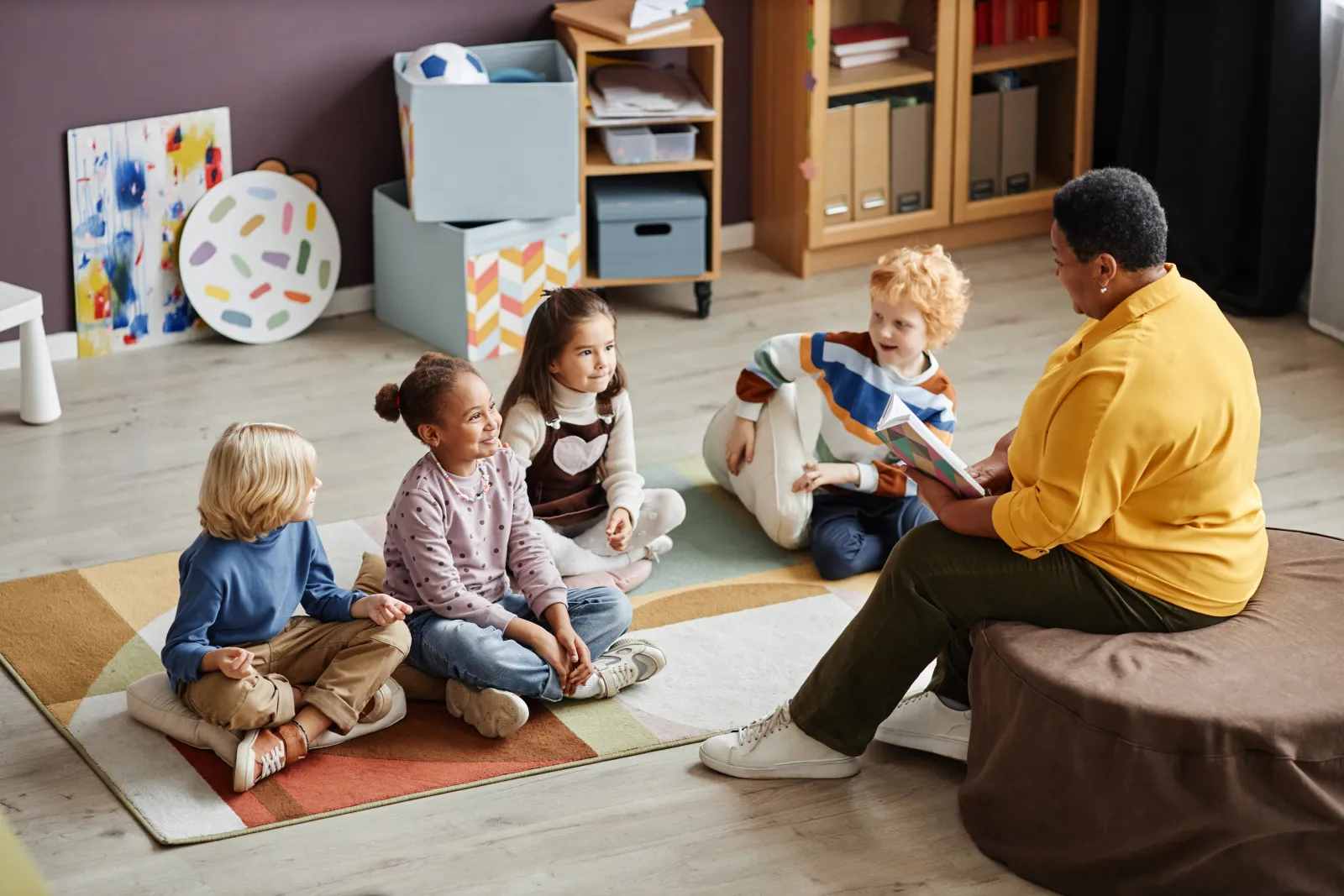 Une adulte lit un livre à quatre jeunes enfants assis sur un tapis.