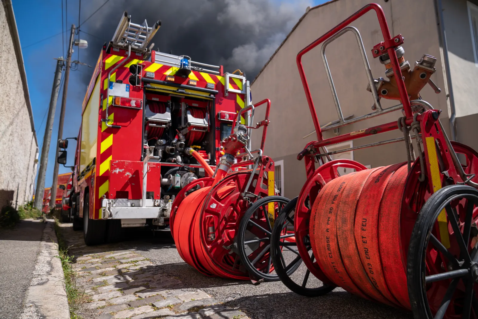 Un engin de pompiers et des dévidoirs de tuyaux sont déployés près d’un bâtiment enfumé.