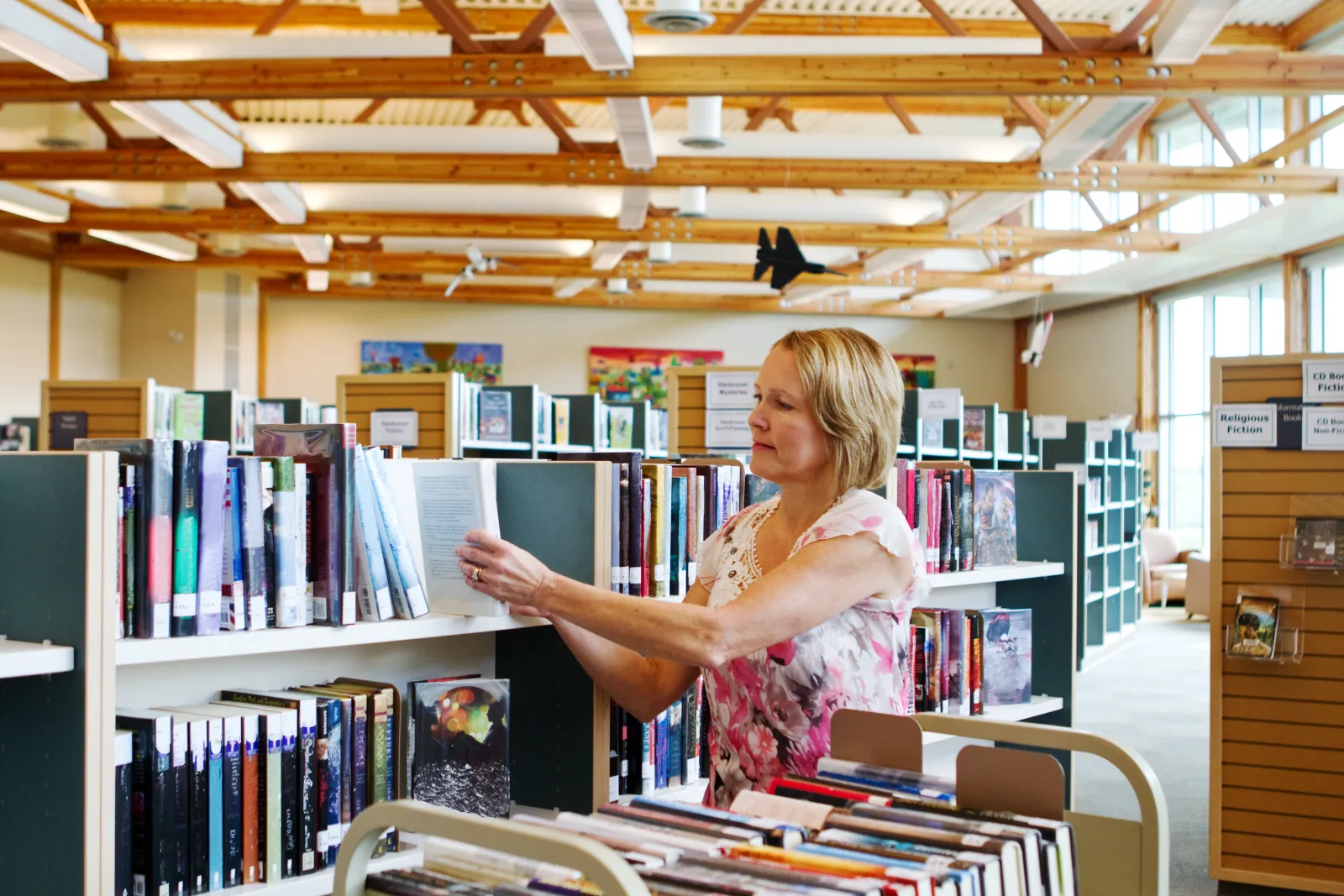 Une bibliothécaire range des livres en rayon, à côté d’un chariot de documents.